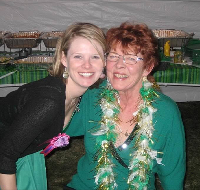 Two women posing for a photo together, in front of a buffet table. One woman, with blonde hair, in a green dress and a black sweater. The other woman with red hair and glasses, in a green shirt, with a feather necklace on.