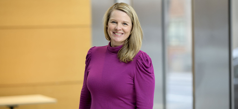 A woman with blonde hair in a purple turtleneck stands, smiling, against a wood and silver backdrop.