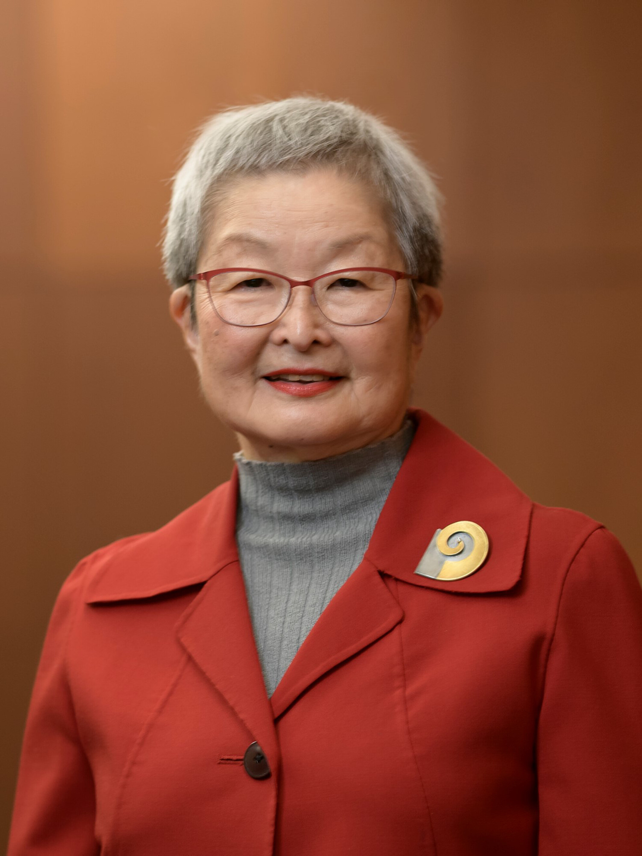 A woman with short gray hair in a gray mock turtleneck sweater and red blazer with a gold brooch stands, smiling, against a wood backdrop. 