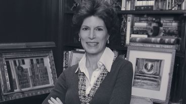 Woman smiling with arms crossed, standing in front of a bookshelf with framed pictures.