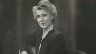 A black-and-white portrait of a woman with curly hair, seated and wearing a dark suit.