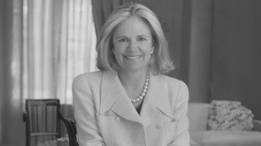 Smiling woman in a white coat with pearls, seated in a cozy room. Black and white photograph.