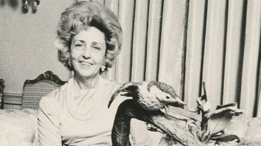 Smiling woman with styled hair, pearl necklace, and bird sculpture; seated on a sofa.
