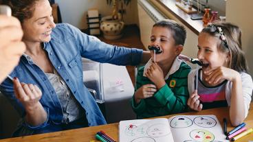 A woman smiles at two laughing children who are holding paper mustaches.