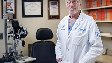 Doctor in a white coat standing in an exam room with ophthalmology equipment.