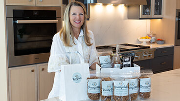 Granny-O-La founder Jennifer stands in a kitchen with a display of Granny-O-La products in front of her. 